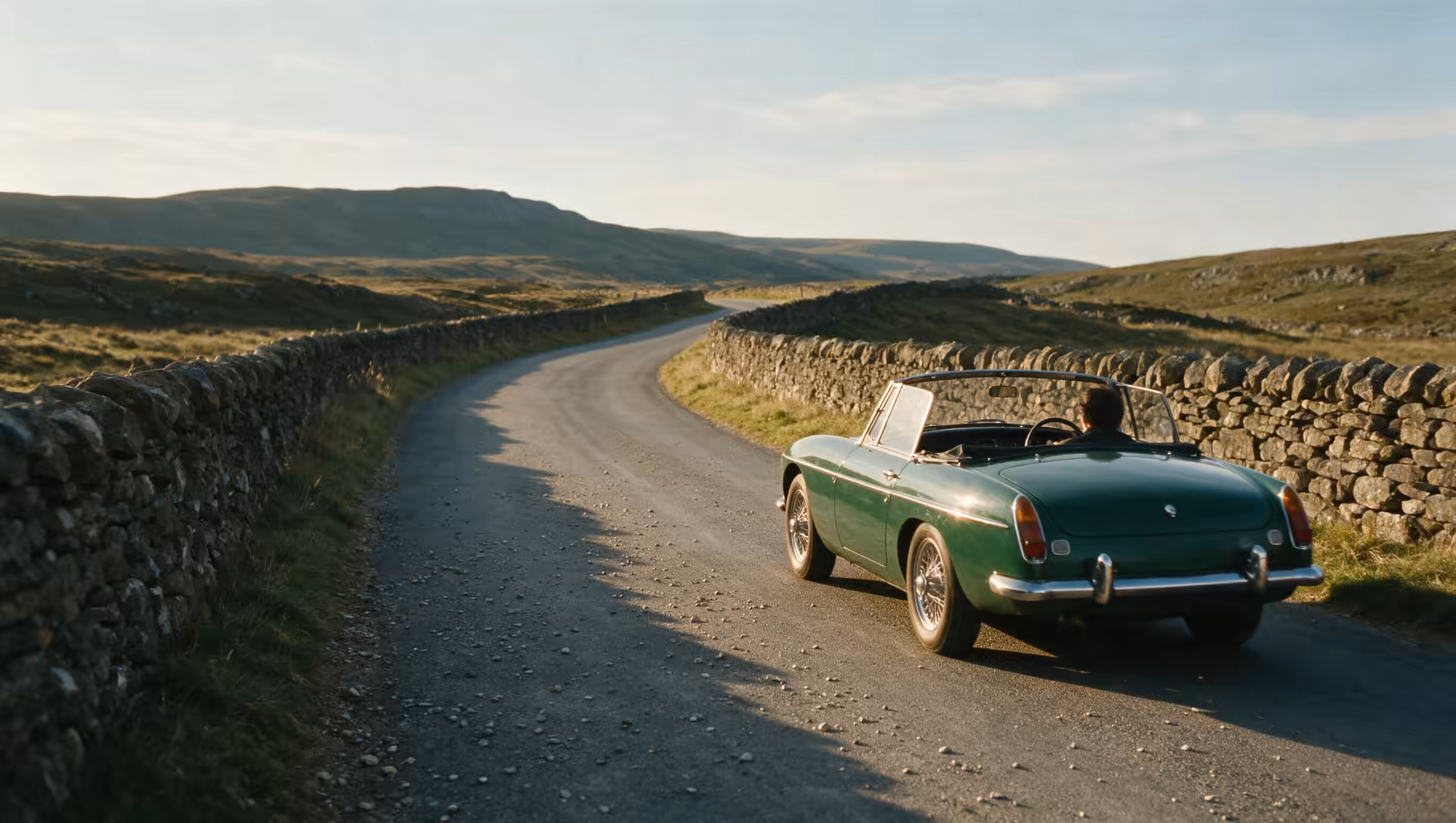 A green classic roadster on a narrow walled road in the Dales, low evening sun